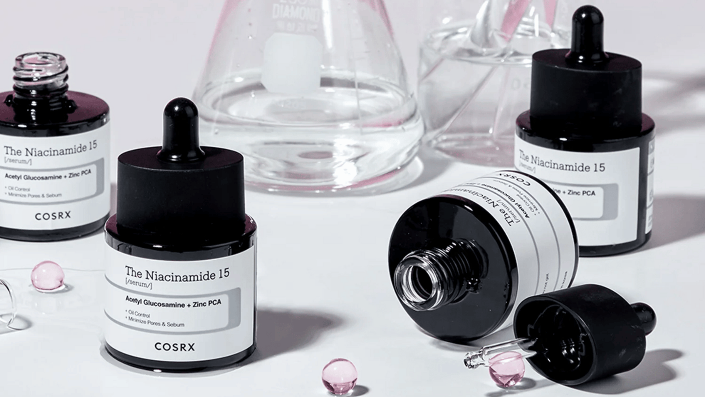 COSRX The Niacinamide 15 Serum med acetylglukosamin och zink PCA, designad för att kontrollera olja och minimera porer. Flaskorna är omgivna av pipetter och droppar på en vit yta, med glasbehållare i bakgrunden som förstärker den vetenskapliga känslan.