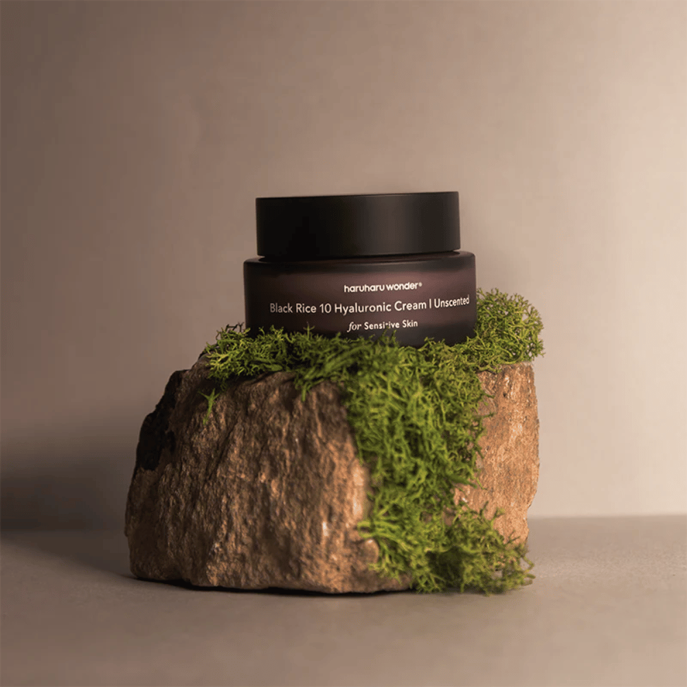 Bilden visar en burk hudkräm märkt "haruharu wonder Black Rice 10 Hyaluronic Cream Unscented for Sensitive Skin". Burken är svart och står på en stor sten täckt med ljusgrön mossa, vilket skapar en naturlig och jordnära känsla.