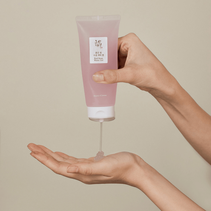 Bilden visar en person som håller en tub av "Red Bean Water Gel" från Beauty of Joseon och klämmer ut en genomskinlig gel i sin hand. Produkten ser ut att vara avsedd för hudvård och har en attraktiv, mjuk rosa förpackning.