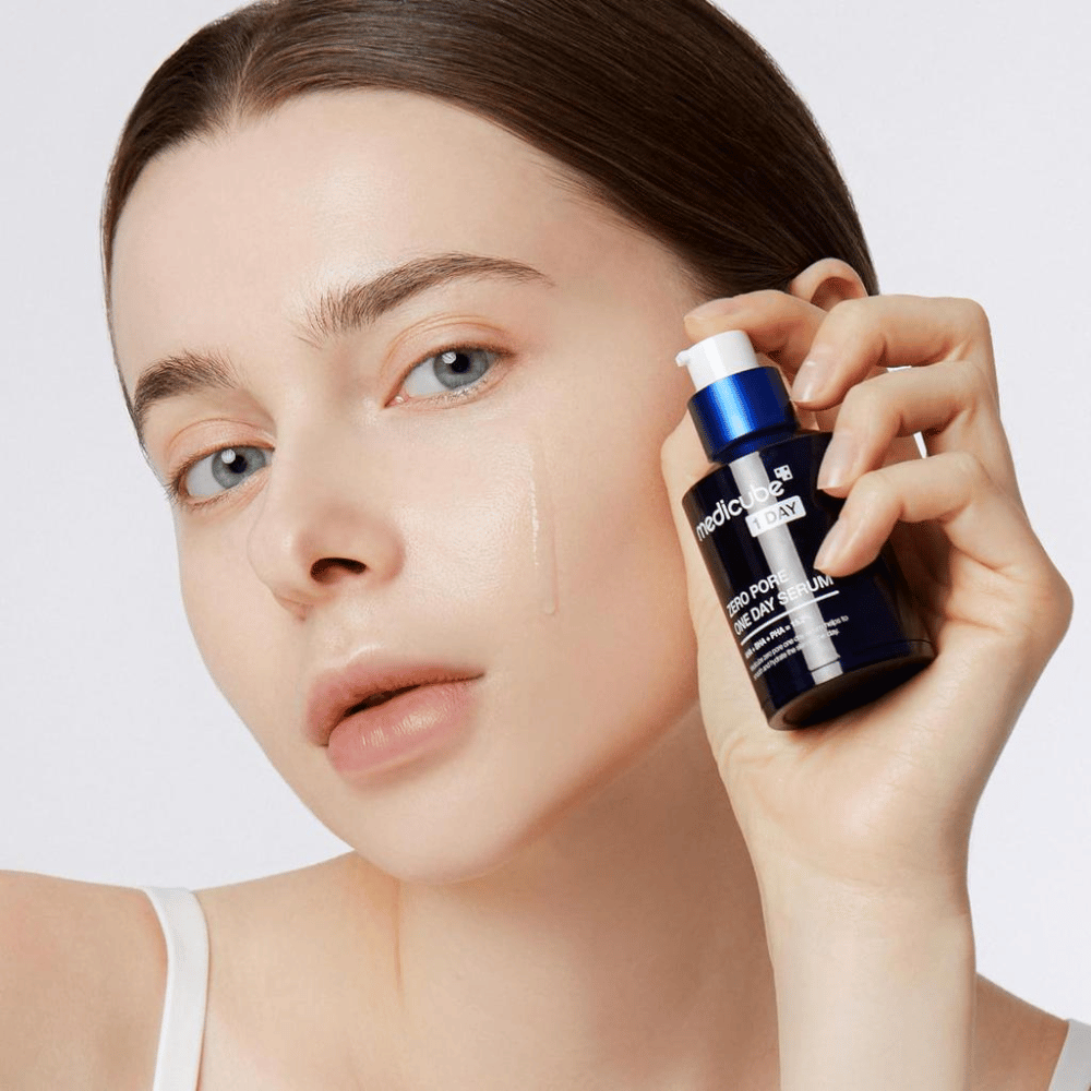 Kvinna med naturlig look håller Medicube Zero Pore One Day Serum och applicerar en droppe på sin kind. Produkten är designad för att exfoliera och förbättra hudens struktur med AHA, BHA och PHA. Perfekt för en slätare och klarare hy.